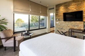A modern hotel room with a large bed, a flat-screen TV mounted on a stone wall, a small round table, and a seating area with a chair and a bench. - Ananti Resort & Spa (Trikala)