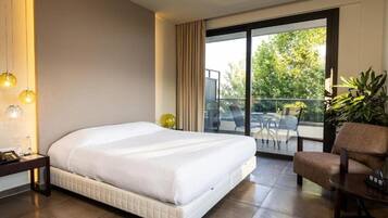 Habitación de hotel con una cama grande, una mesita, una silla, un balcón con puerta de vidrio y una planta en maceta.