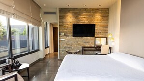 A hotel room with a large bed, a flat-screen TV mounted on a stone wall, a desk with a chair, and a window with curtains. - Ananti Resort & Spa (Trikala)