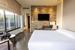 A hotel room with a large bed, a flat-screen TV mounted on a stone wall, a desk with a chair, and a window with curtains. - Ananti Resort & Spa (Trikala)