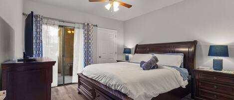 A bedroom with a bed, a nightstand, a dresser, a ceiling fan, and a door leading to a bathroom.