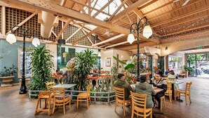 A spacious indoor dining area with wooden tables and chairs, surrounded by potted plants and a high ceiling with exposed beams.