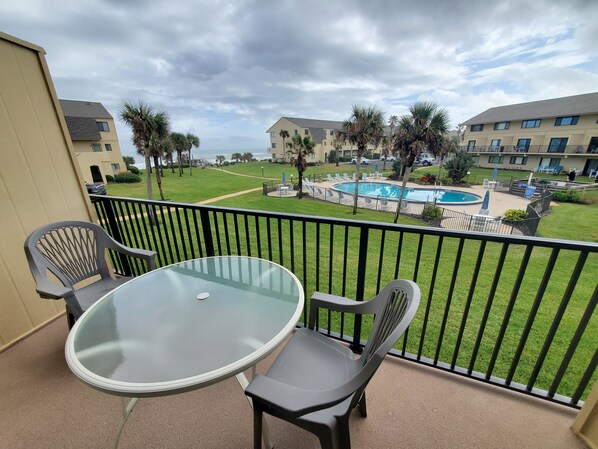 A balcony with a glass table and two chairs, overlooking a pool area with palm trees and a building in the background.