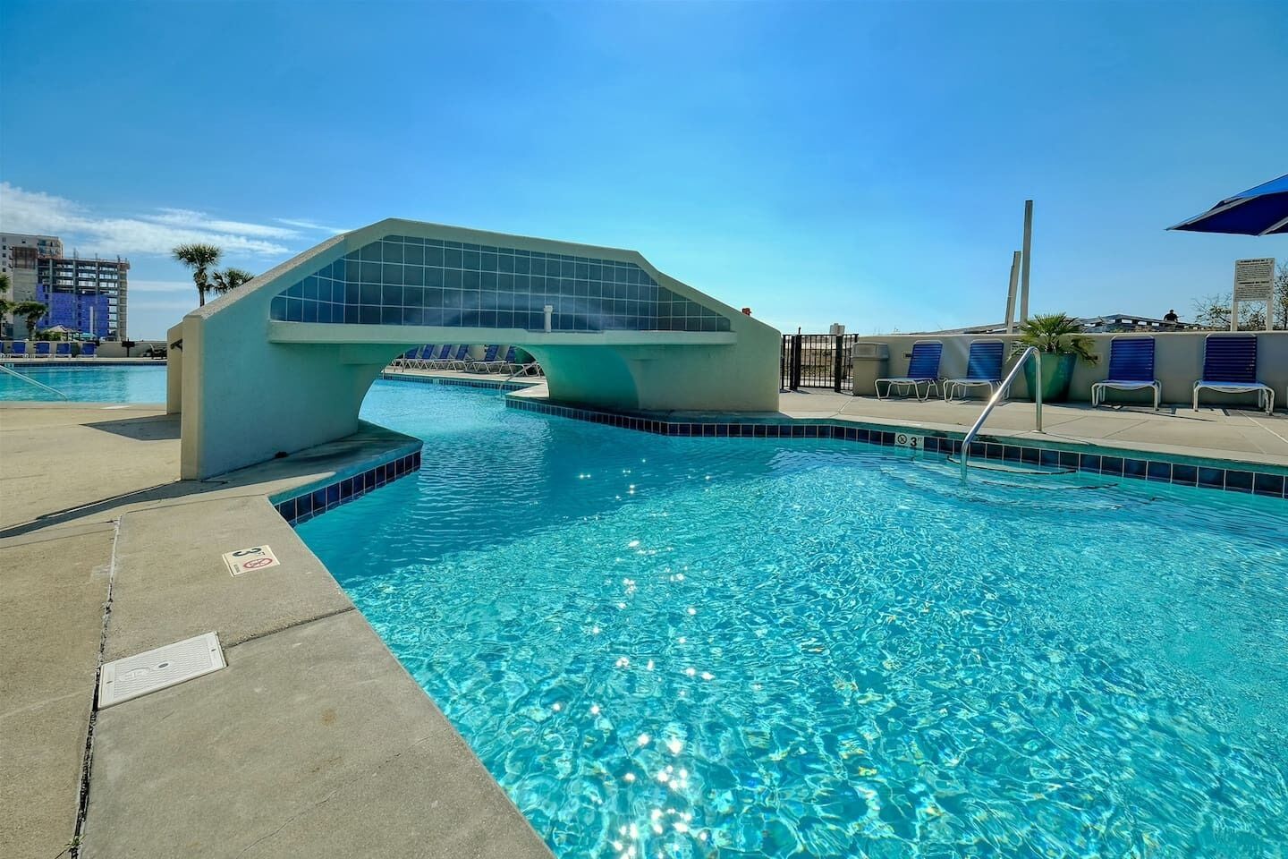 Ein moderner Poolbereich mit einer markanten, bogenförmigen Glasbrücke, klarem, blauem Wasser und Liegestühlen.