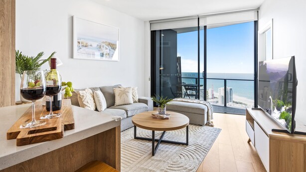 A modern living room with a sofa, coffee table, and a large TV. There are wine glasses and a bottle on a wooden board. The room has large windows with a view of the city.