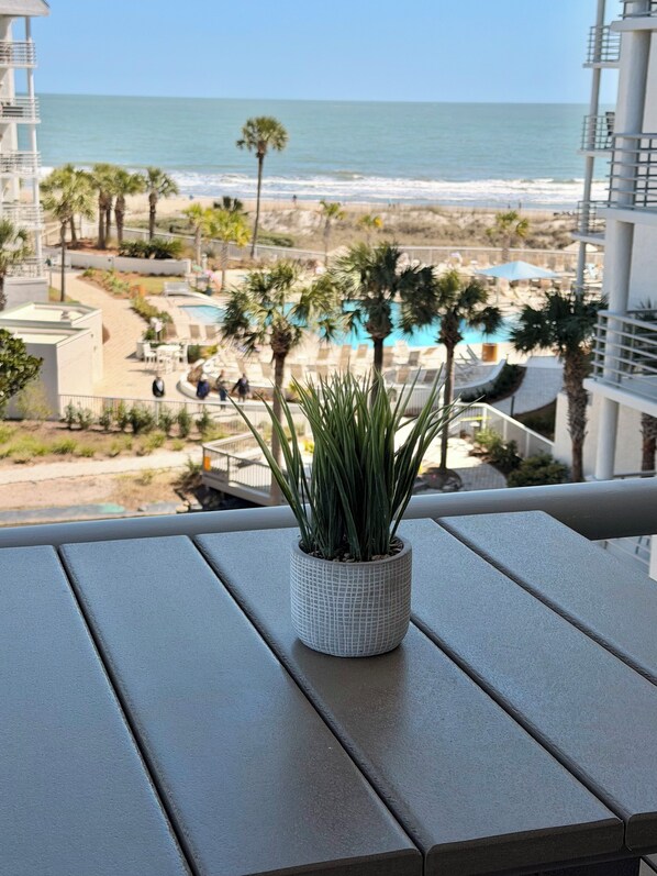 Ein Balkon mit einem Blumentopf, mit Blick auf einen Strand mit Palmen und einen Poolbereich.