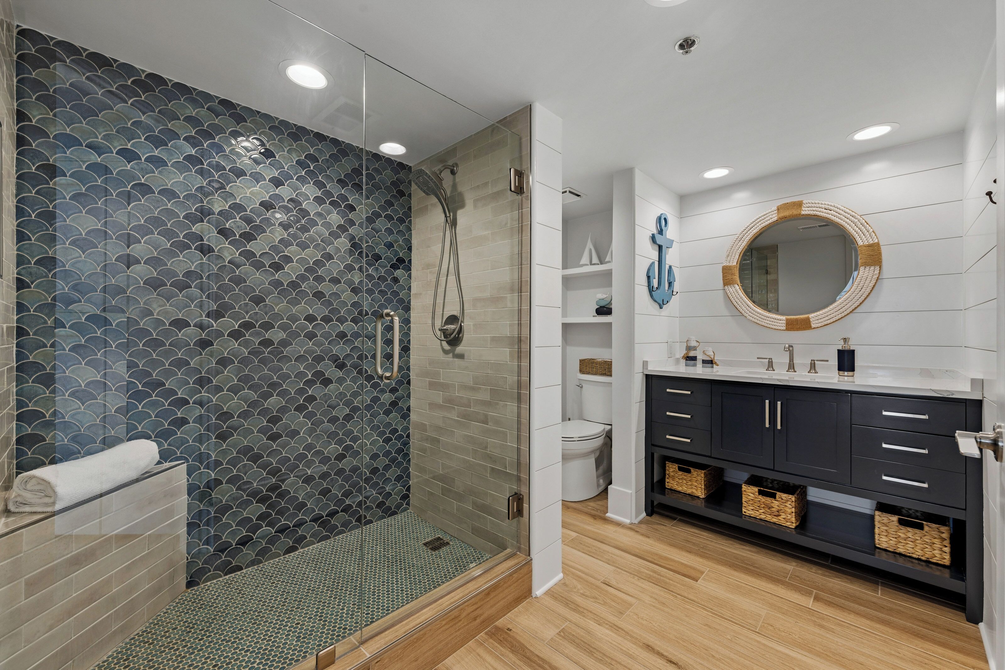 Une salle de bain moderne avec une douche à parois de verre, des murs recouverts de carreaux blancs, un plancher en bois et un meuble-lavabo foncé doté de tiroirs et de paniers.