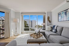 A modern living room with a grey sofa, a wooden coffee table with candles, and a large window offering a view of buildings and trees.