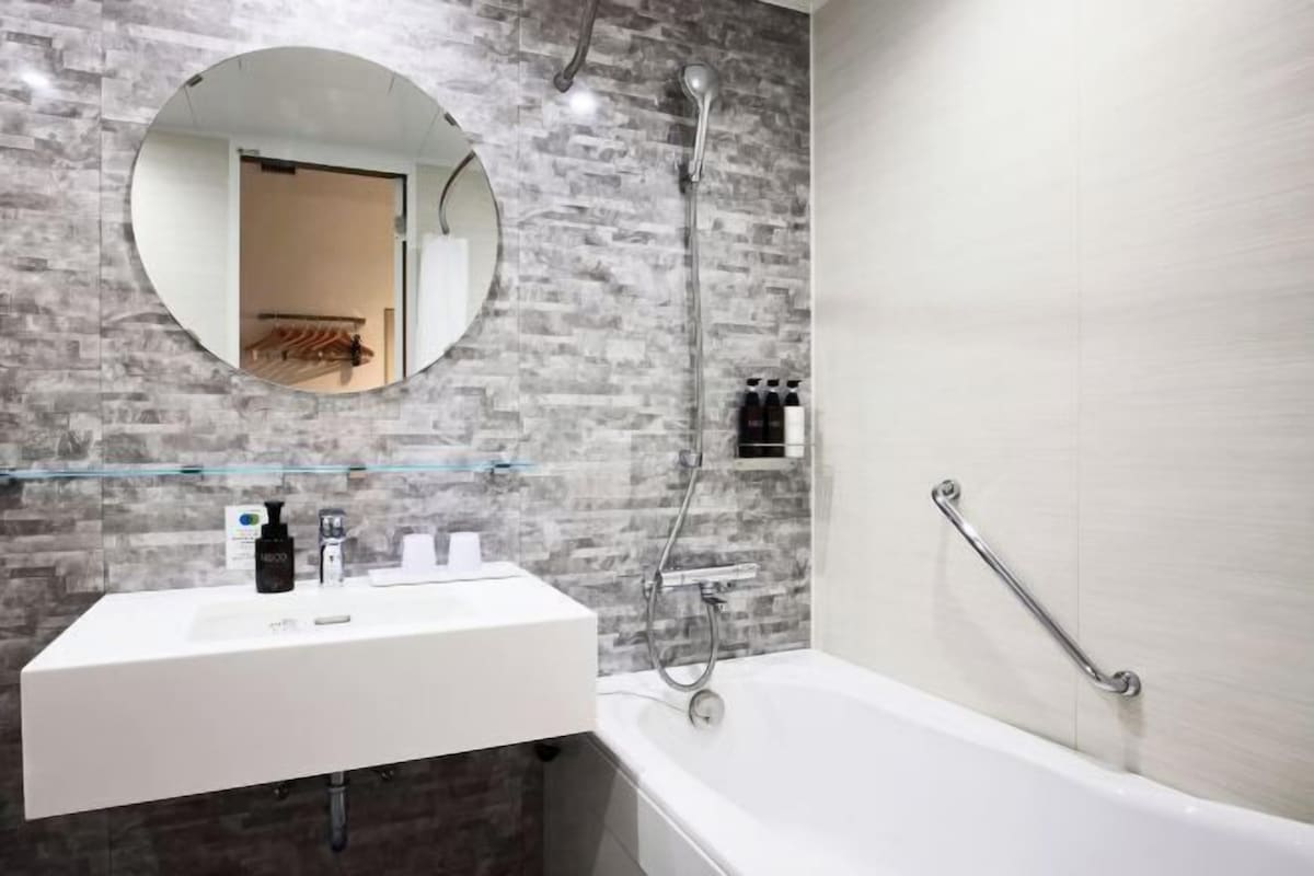 A modern bathroom with a white sink, a round mirror, a shower, and a grab bar.