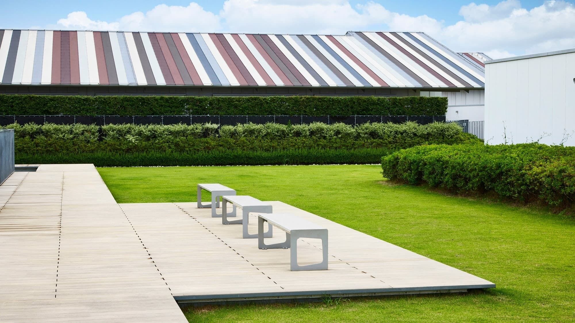 Un edificio moderno con techo a rayas, una terraza de madera con bancos y un césped bien cuidado.