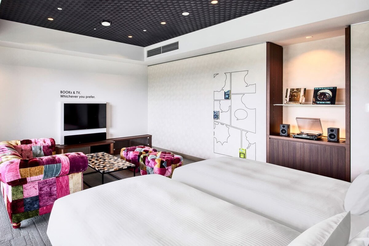 A hotel room with a bed, a TV, a desk, a chair, and a wall with a comic strip design.
