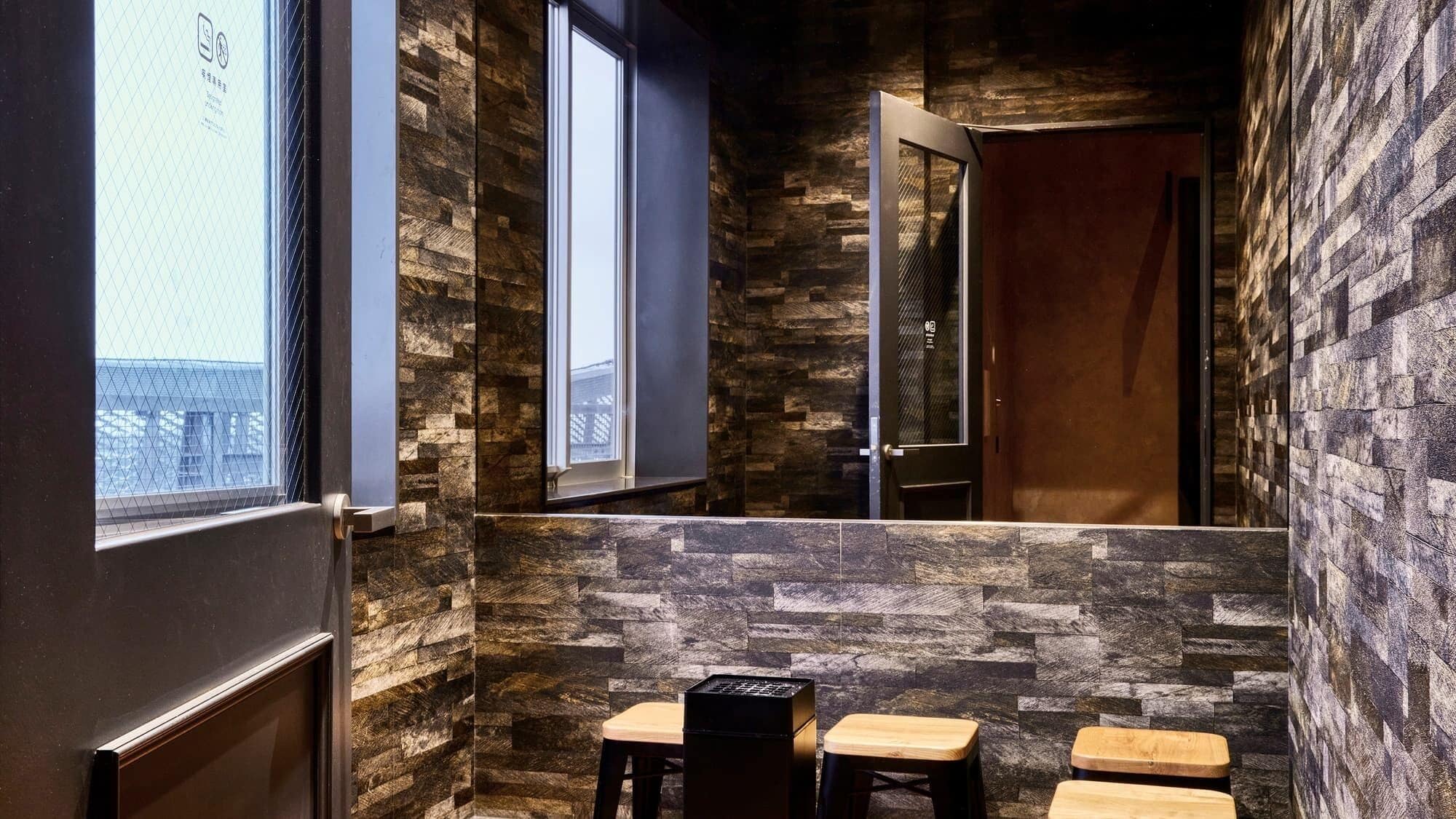 A narrow hallway with stone walls, a door, and a window. There are three wooden stools and a black object on the floor.