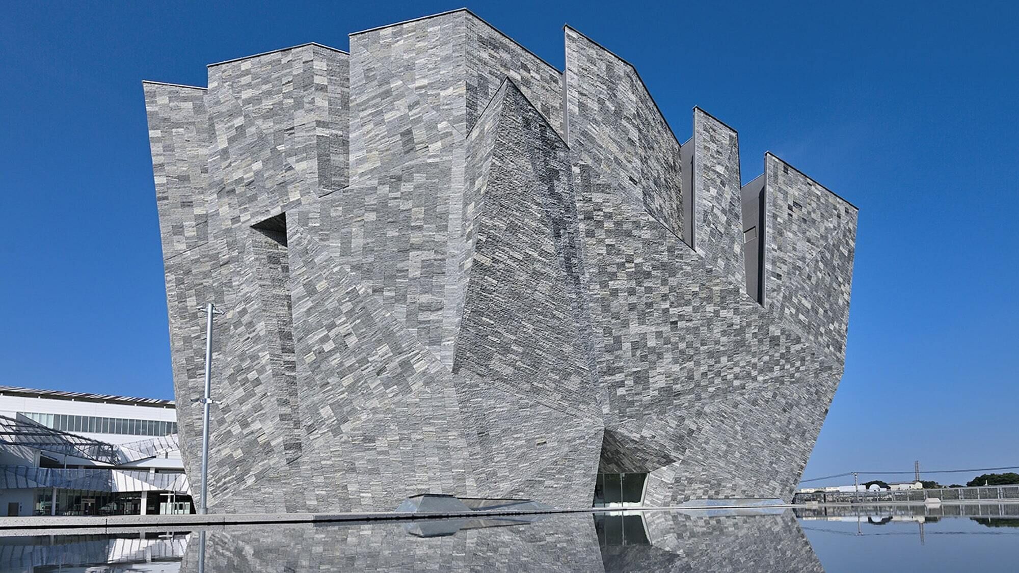 Un edificio moderno con fachada geométrica singular, una superficie acuática reflectante en primer plano y un cielo azul despejado al fondo.