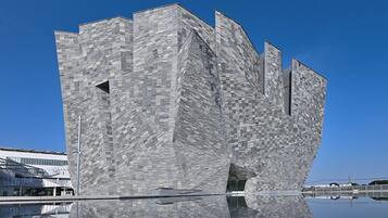 Un edificio moderno con fachada geométrica singular, una superficie acuática reflectante en primer plano y un cielo azul despejado al fondo.
