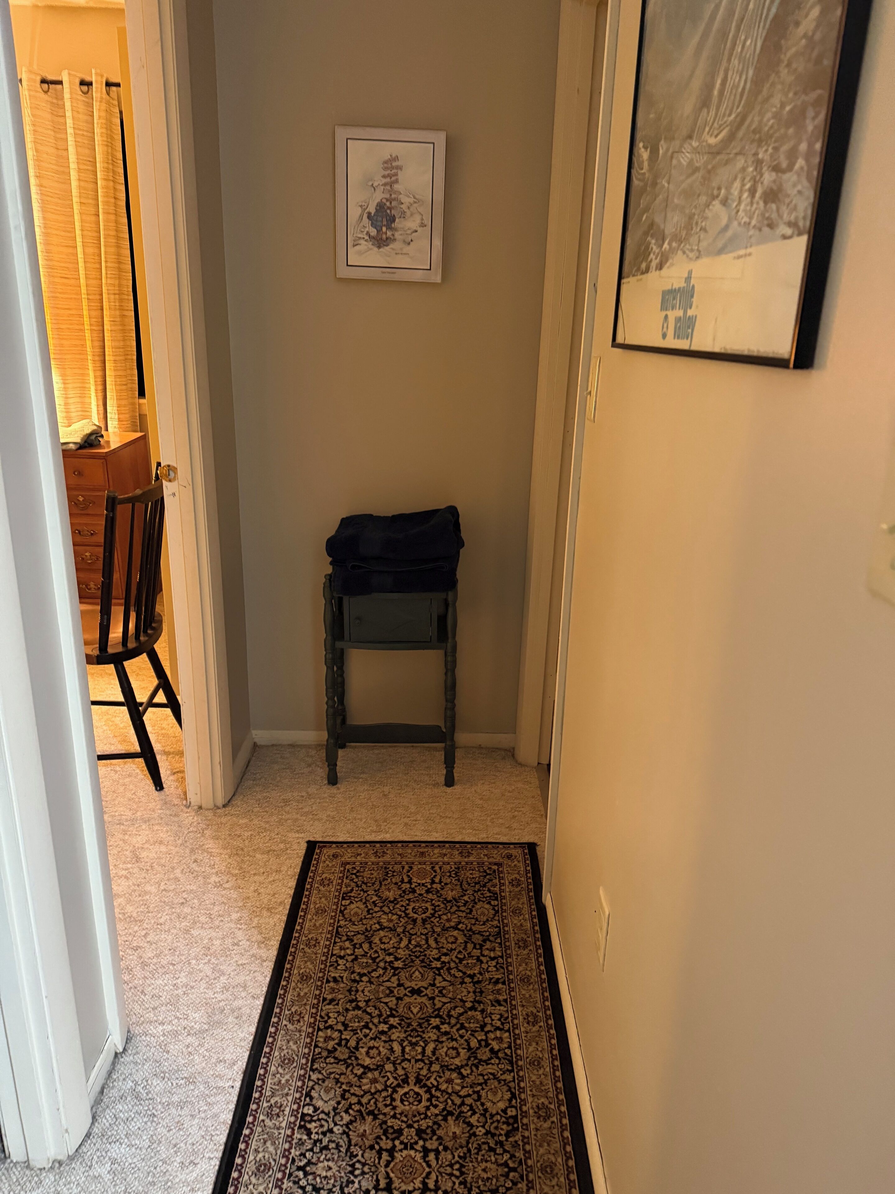 Een hal met een kamerbreed tapijt, een klein tafeltje met een opgevouwen voorwerp, en een lijst met een afbeelding aan de muur.