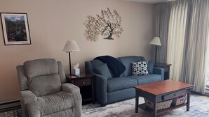 Una sala de estar con un sillón gris, un sofá azul, una mesa de centro de madera y un elemento decorativo de arte mural con un árbol.