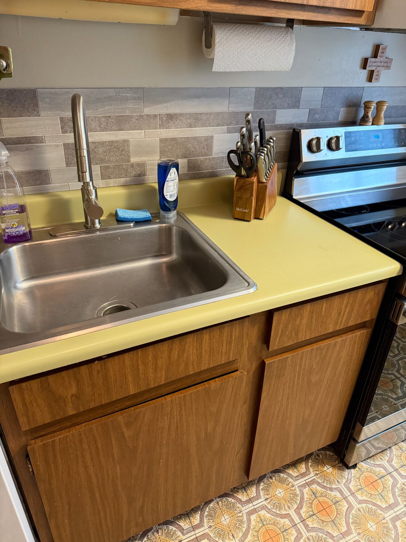 A kitchen with a stainless steel sink, wooden cabinets, a yellow countertop, a patterned backsplash, and a stove.