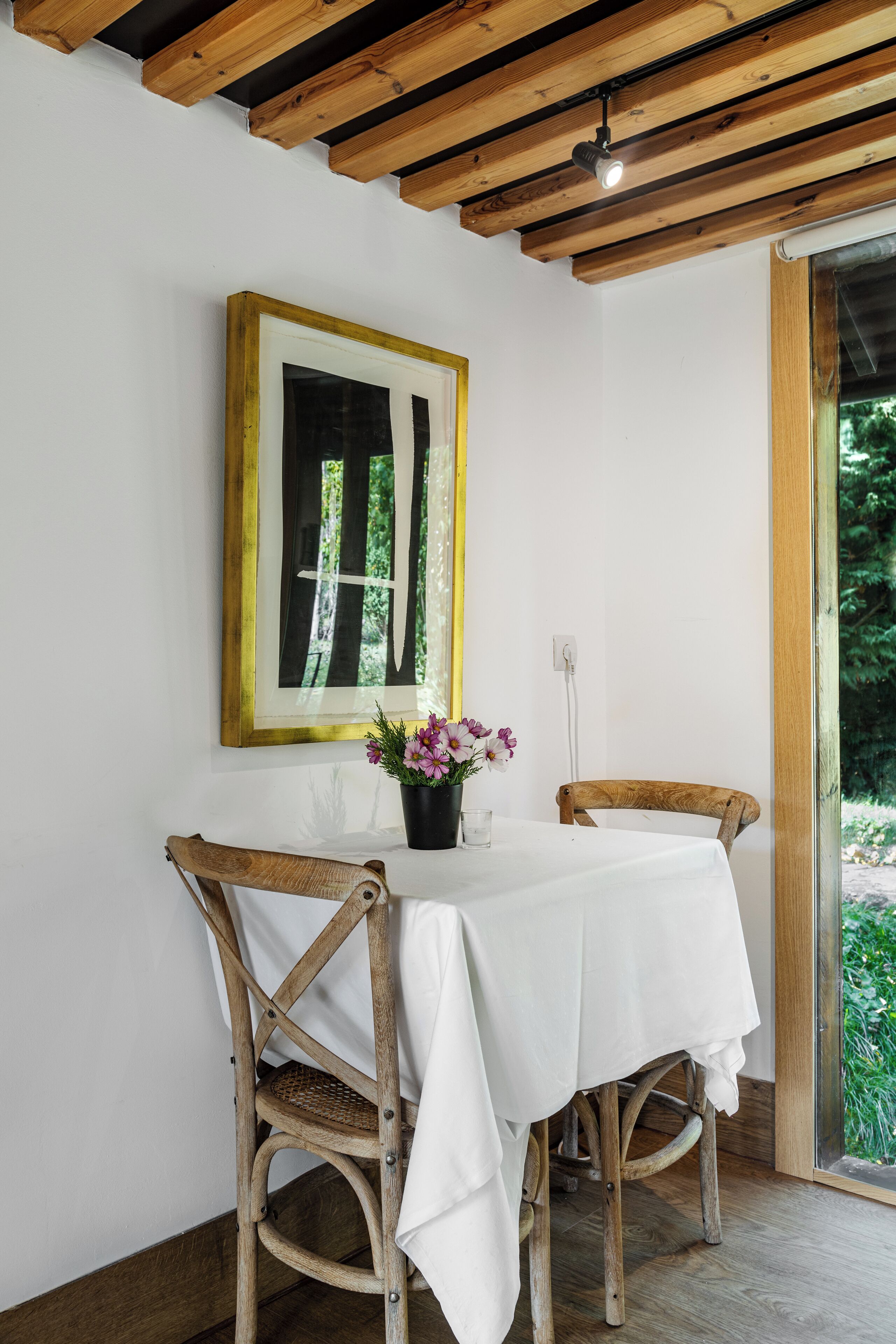 A dining area with a table set for two, wooden chairs, a framed mirror, and a potted plant.