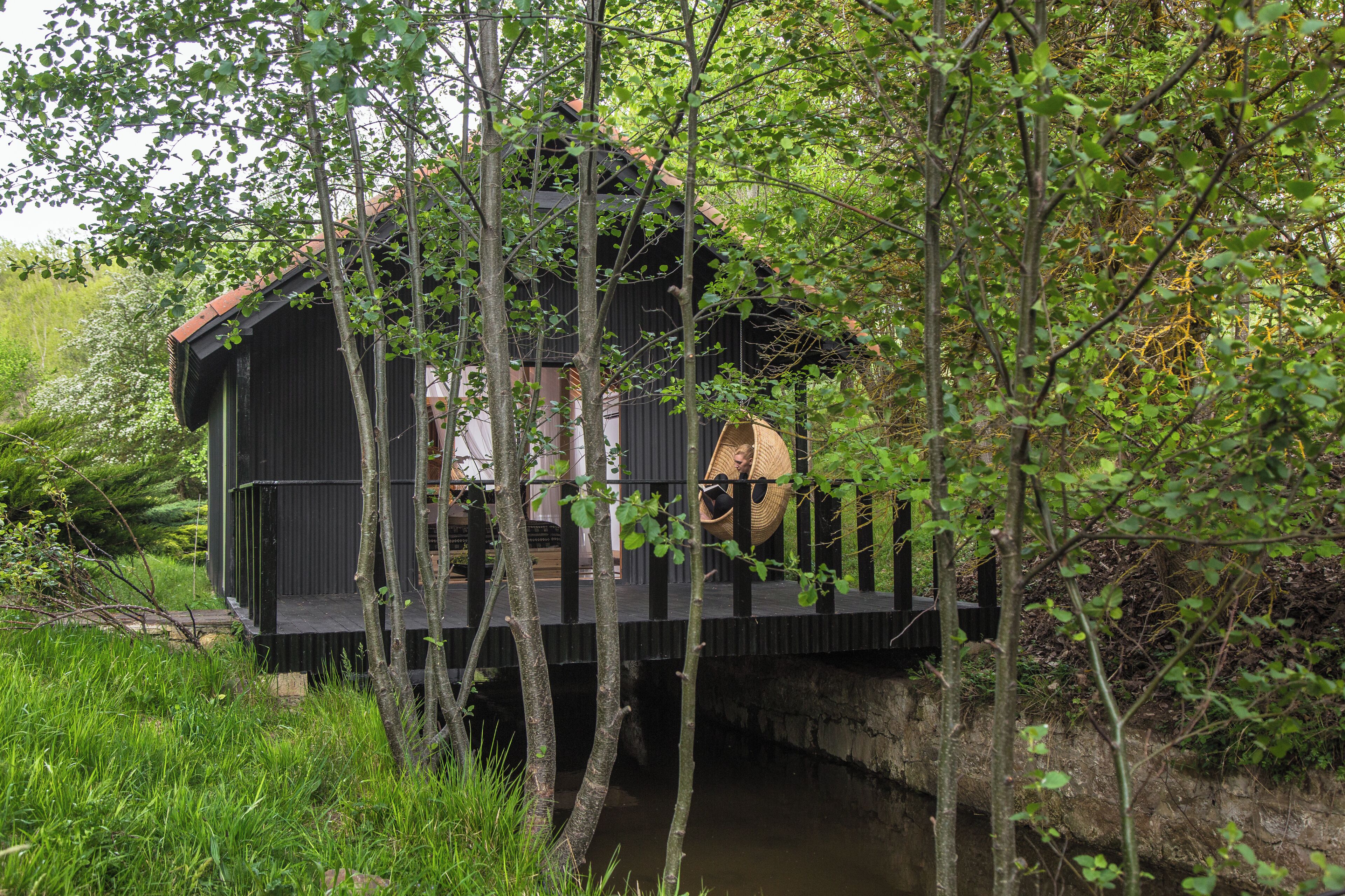 A wooden cabin with a balcony, surrounded by lush greenery and a small stream.