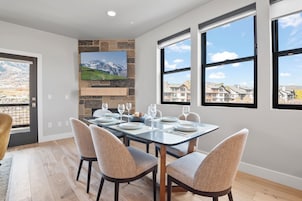 Un área de comedor con pared de piedra, un televisor de pantalla plana y ventanas amplias que ofrecen vistas a un barrio residencial.