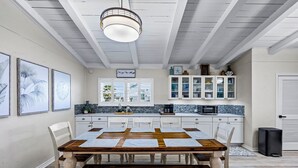 Un'area pranzo con tavolo e sedie in legno, una cucina con mobili bianchi e zoccolo in piastrelle, e una ventola a soffitto.