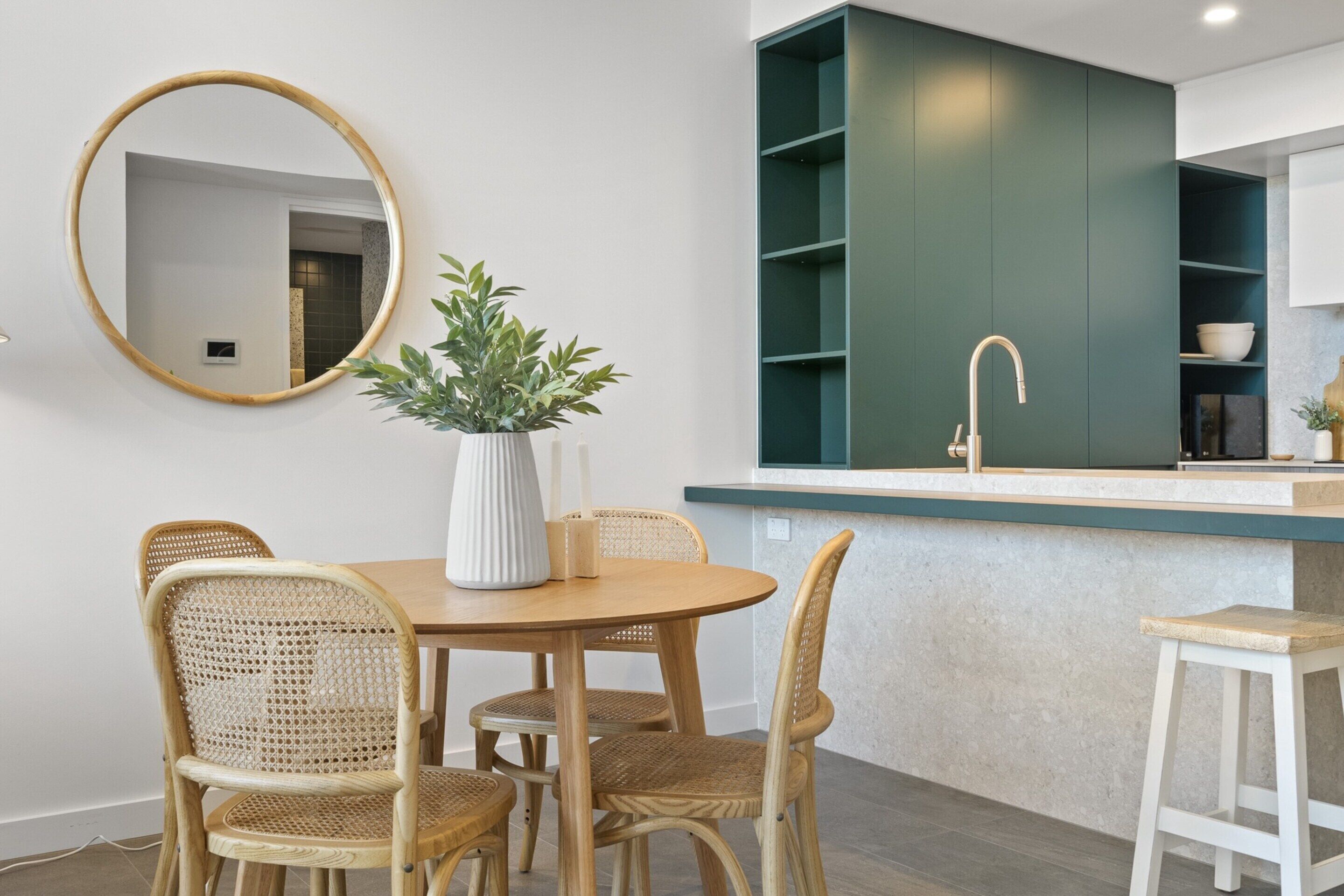 A modern kitchen with a round dining table, wicker chairs, a white vase with green plants, and a green cabinet with open shelves.