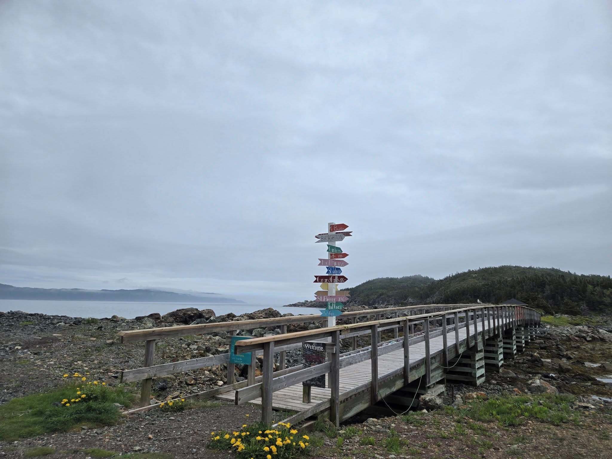 A wooden pier leading to a rocky shore with directional signs pointing to various destinations.