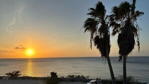 A beach scene with a sunset, palm trees, and a person sitting on a bench. - Uncle Bob`s Reef: Oceanfront Paradise – 1 Bedroom, 2 Bath Townhome (Sabana Westpunt)