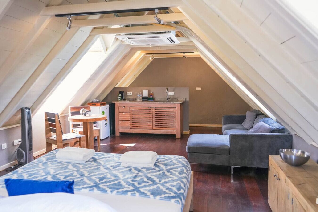 Une chambre à coucher compacte, avec un toit en pente, comprenant un lit, une table de chevet et un petit bureau avec une chaise. On aperçoit un coin cuisine au fond, et un canapé gris est placé contre le mur.