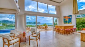 A spacious room with large windows offering a view of the sea and hills. There is a bar area with stools and a table with chairs.