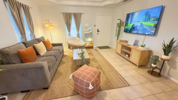 Una sala de estar con sofá, sillón, mesa de centro, reposapiés, televisor y un mueble de madera con plantas.