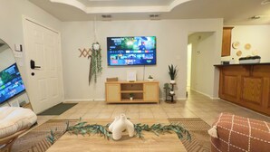 Una sala de estar con un televisor de pantalla plana montado en la pared, un mueble de madera para entretenimiento, una mesa de centro con jarrones decorativos y una puerta con perchero.