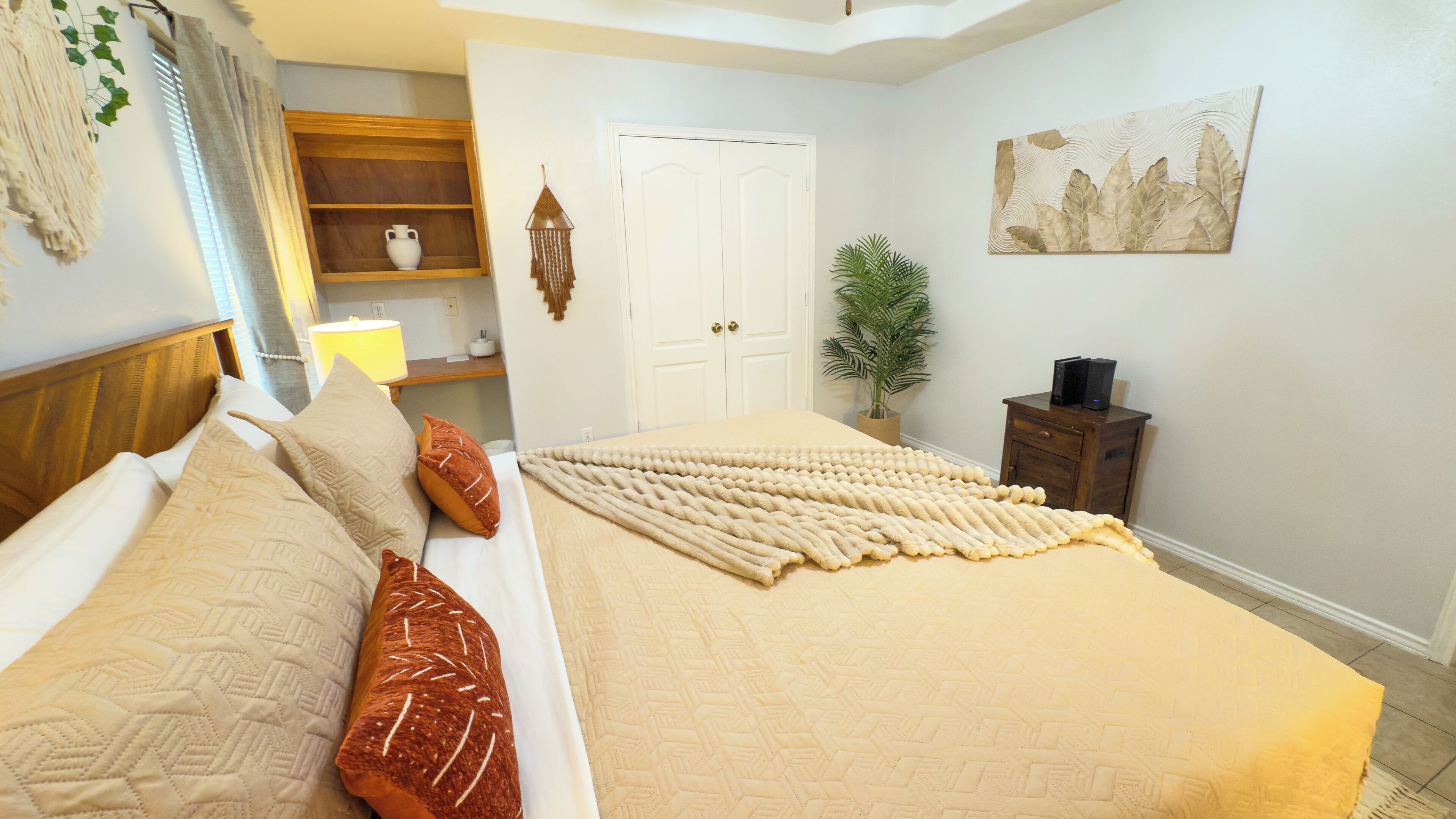 A bedroom with a bed, pillows, a bedside table, a lamp, a plant, a door, and a wall decoration.