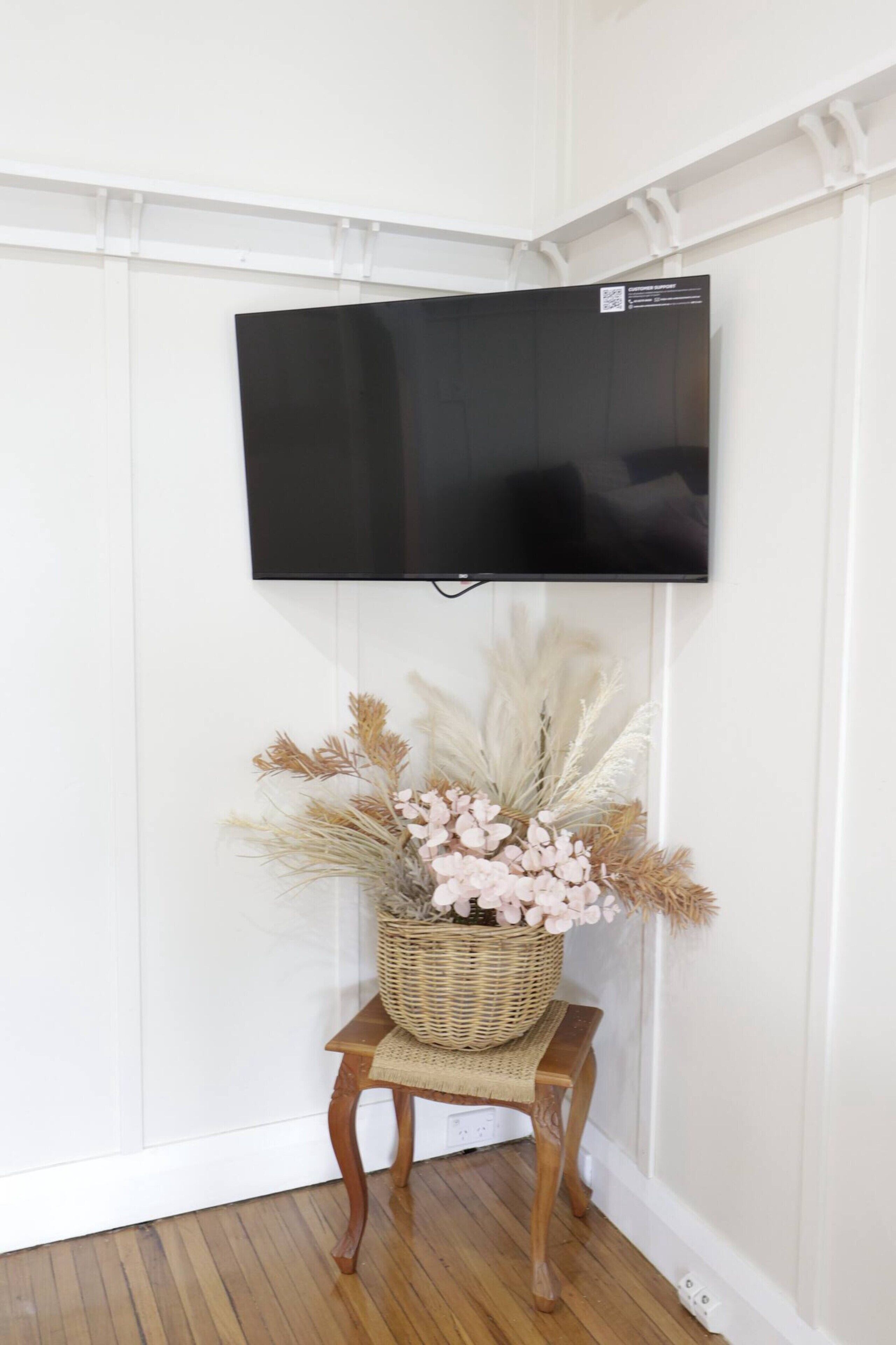 A flat-screen TV mounted on a white wall, a wooden stool with a woven basket underneath, and a decorative arrangement of dried flowers and pampas grass.