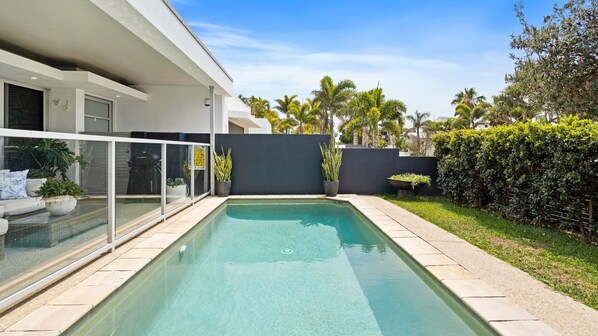 A modern pool area with a clear blue pool, a glass enclosure, and a well-maintained garden with various plants and a hedge.