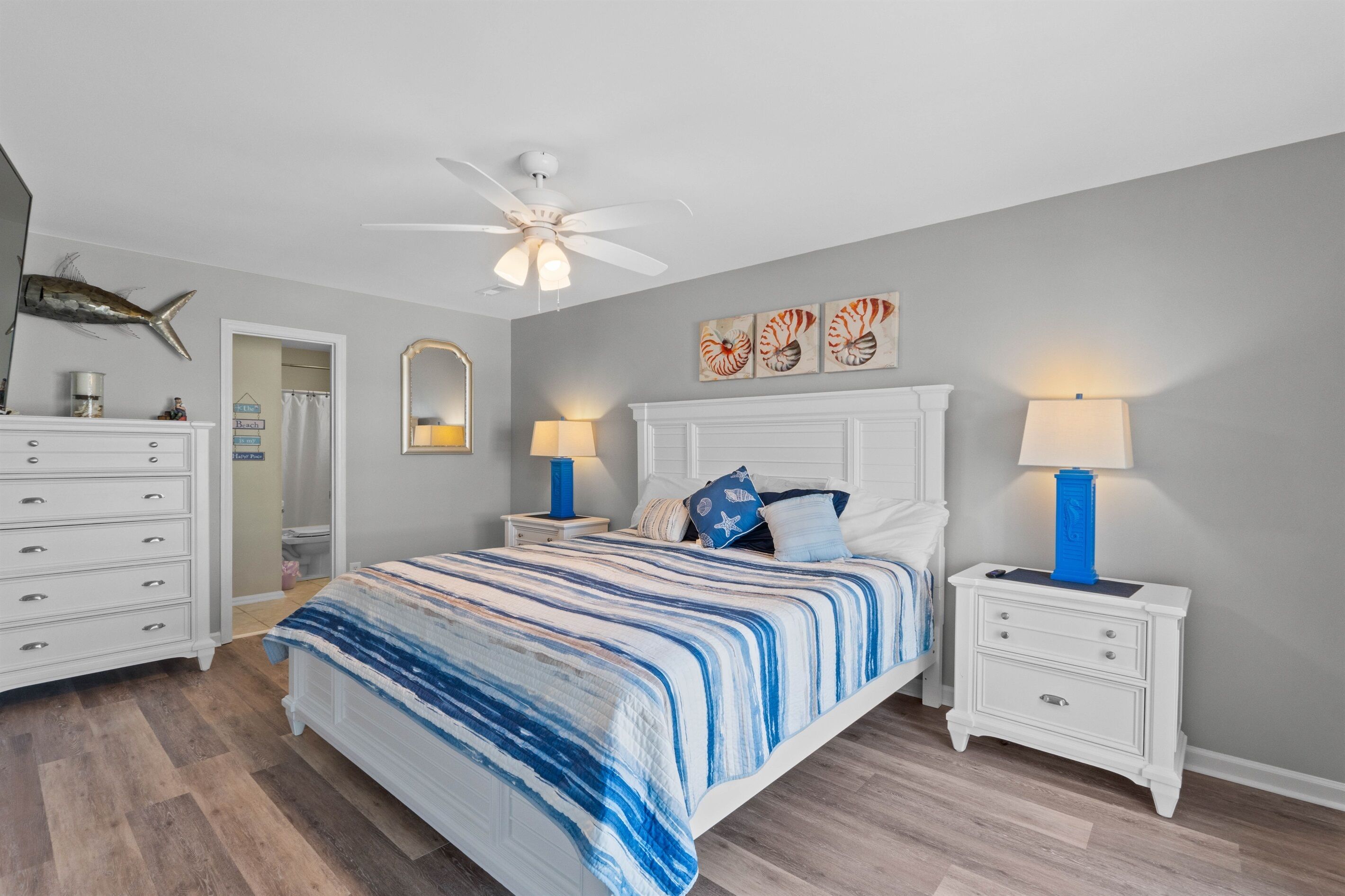 A bedroom with a bed, white nightstands, a ceiling fan, and artwork on the wall.