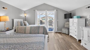 Une chambre avec deux lits, une commode, une télévision et un balcon donnant sur une piscine et des maisons.