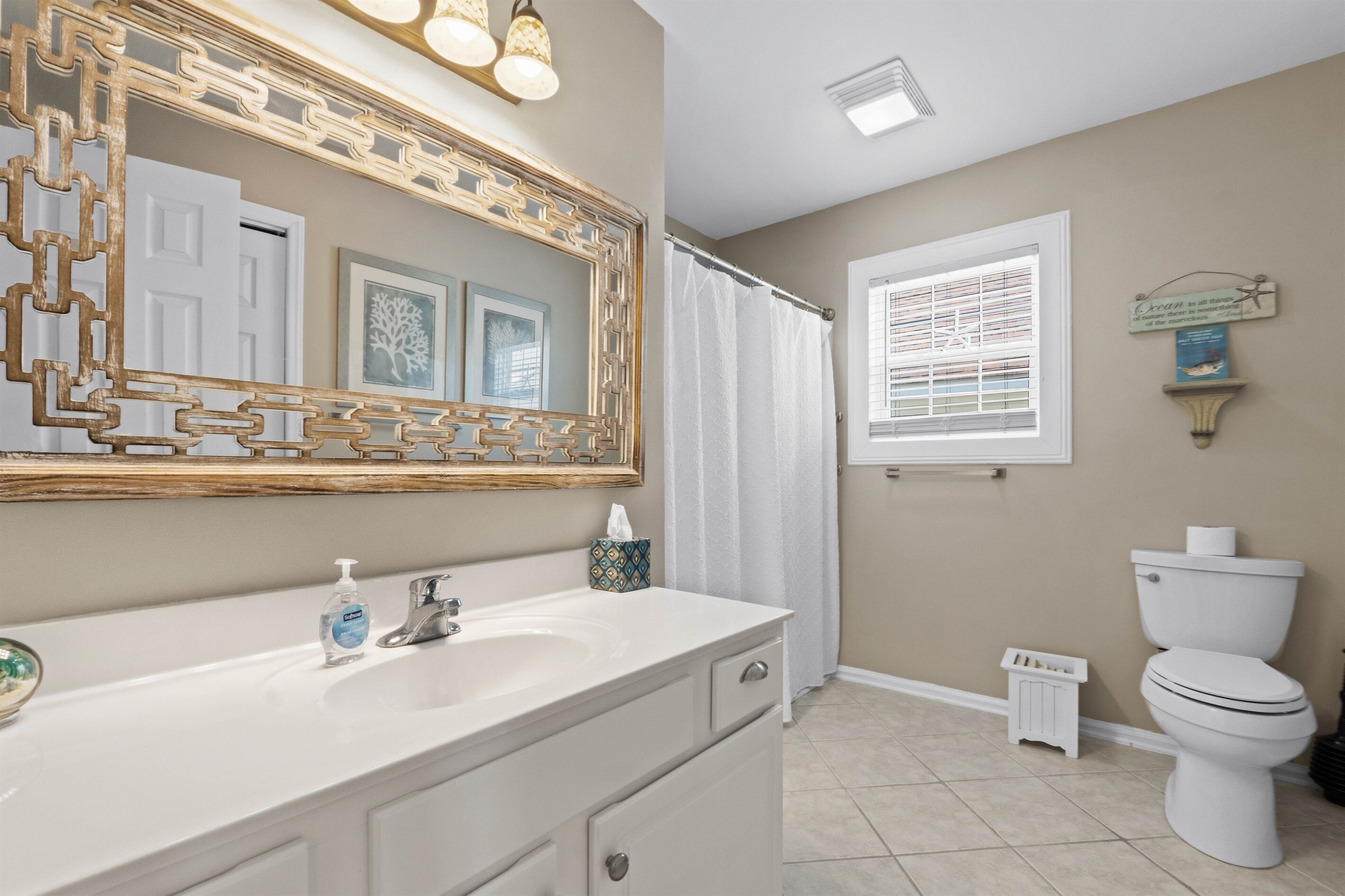 A bathroom with a white sink, a mirror with a decorative frame, a toilet, and a window with blinds.