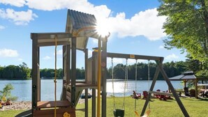 Un terrain de jeux avec un toboggan en bois et des balançoires, situé au bord d’un lac.