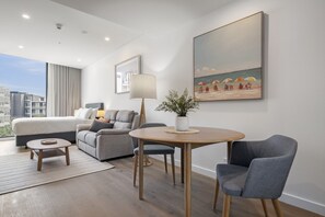 Ein modernes Hotelzimmer mit einem großen Bett, einer Couch, einem Esstisch und einem Stuhl. An der Wand hängt ein Gemälde und das Fenster bietet einen Blick auf Gebäude.