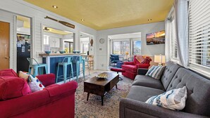 A living room with a red sofa, a grey sofa, and a coffee table. There is a kitchen area with a bar and stools. - Charming dog-friendly cottage w/ Hot Tub, Smart TVs, Community Pool & Pickleball (Avon)