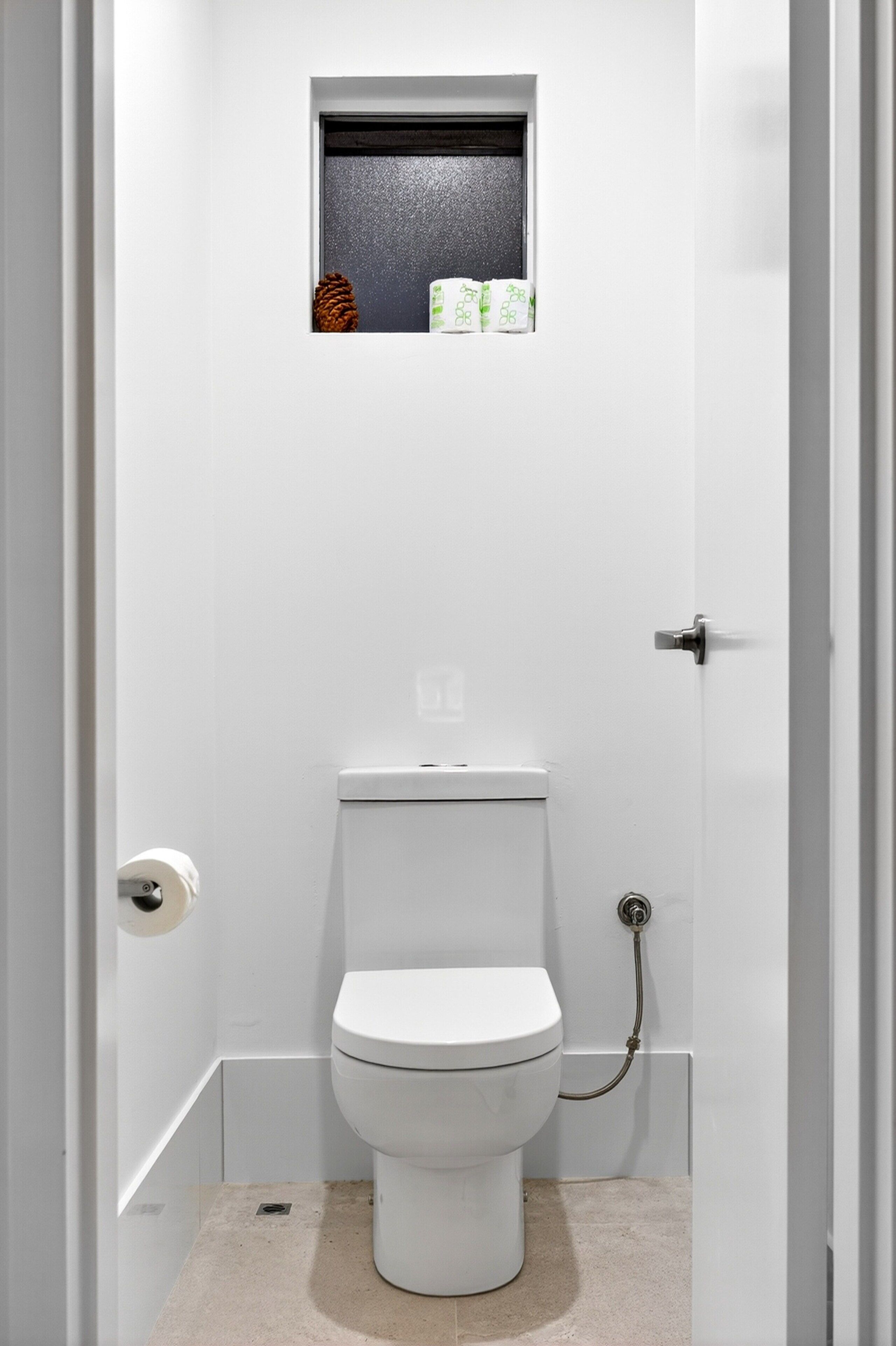 A small, white bathroom with a toilet, a window above it, and a shower area to the right.