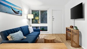 A modern living room with a blue sofa, a wooden coffee table, a painting of a boat on the wall, a TV, and a sliding glass door to the outside. - Bright 2-bed Unit Near Scarborough Beach (Perth)