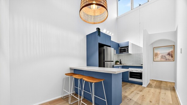 A modern kitchen with blue cabinetry, a central island with stools, a hanging light fixture, and a view of the outdoors. - Bright 2-bed Unit Near Scarborough Beach (Perth)
