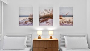 A bedroom with two beds, a wooden nightstand with two lamps, and three beach-themed paintings on the wall. - Bright 2-bed Unit Near Scarborough Beach (Perth)