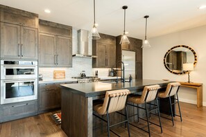 A modern kitchen with wooden cabinets, stainless steel appliances, a central island with bar stools, pendant lighting, and a round mirror on the wall.