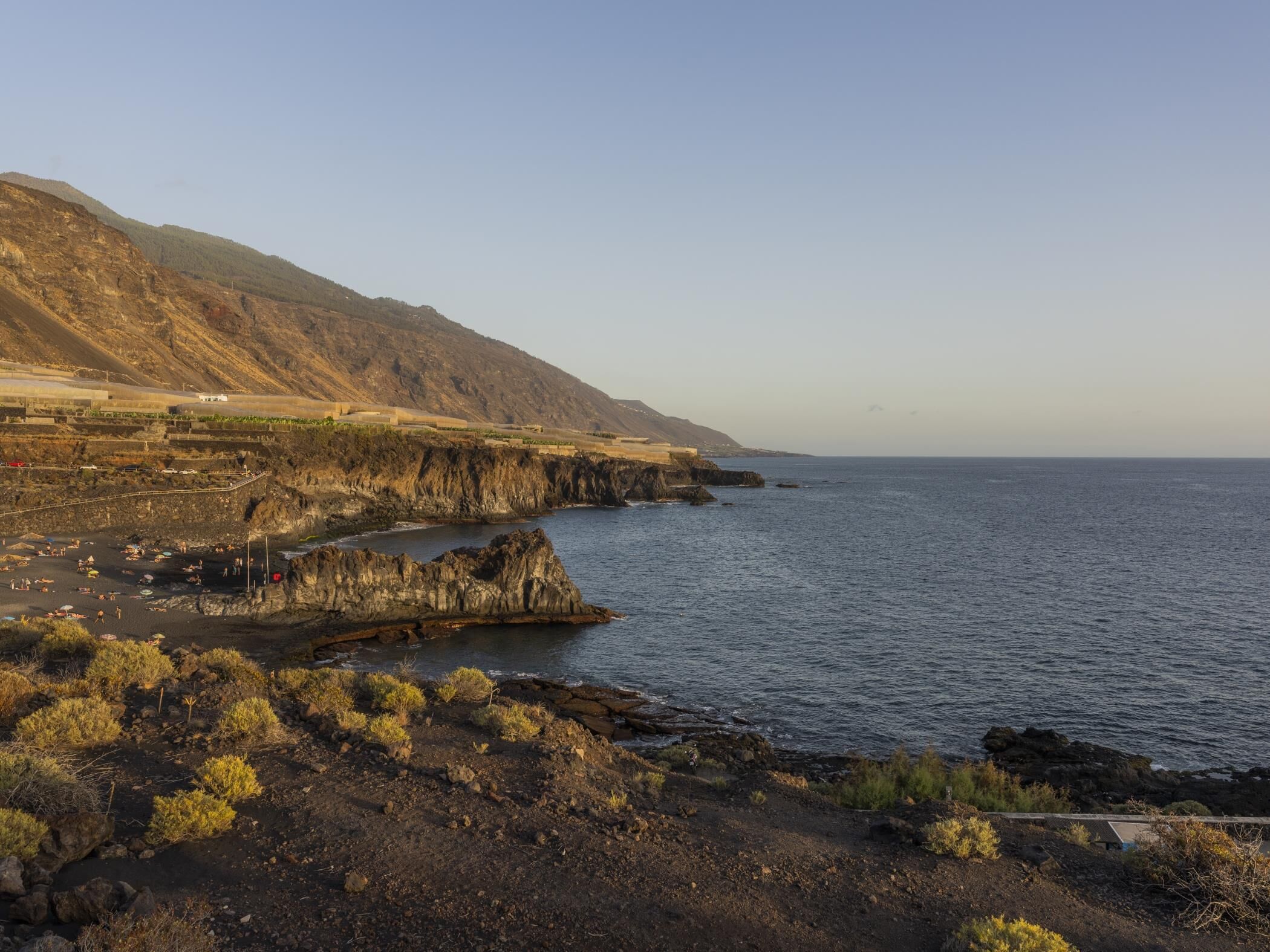 Un paisaje costero con terreno rocoso, una playa y una cadena montañosa distante.