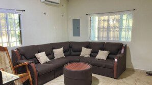 A living room with a dark brown sectional sofa, a wooden ottoman, and a wooden coffee table. There are two windows with metal bars, an air conditioning unit, and a blue wall panel. - Amazing 3 bedroom house with AC in delightful El Salvador in El Truinfo (El Salvador)