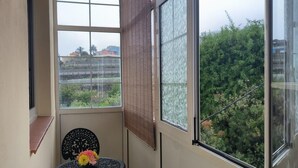 A balcony with a table and chairs, a view of greenery, and a window with a view of buildings.