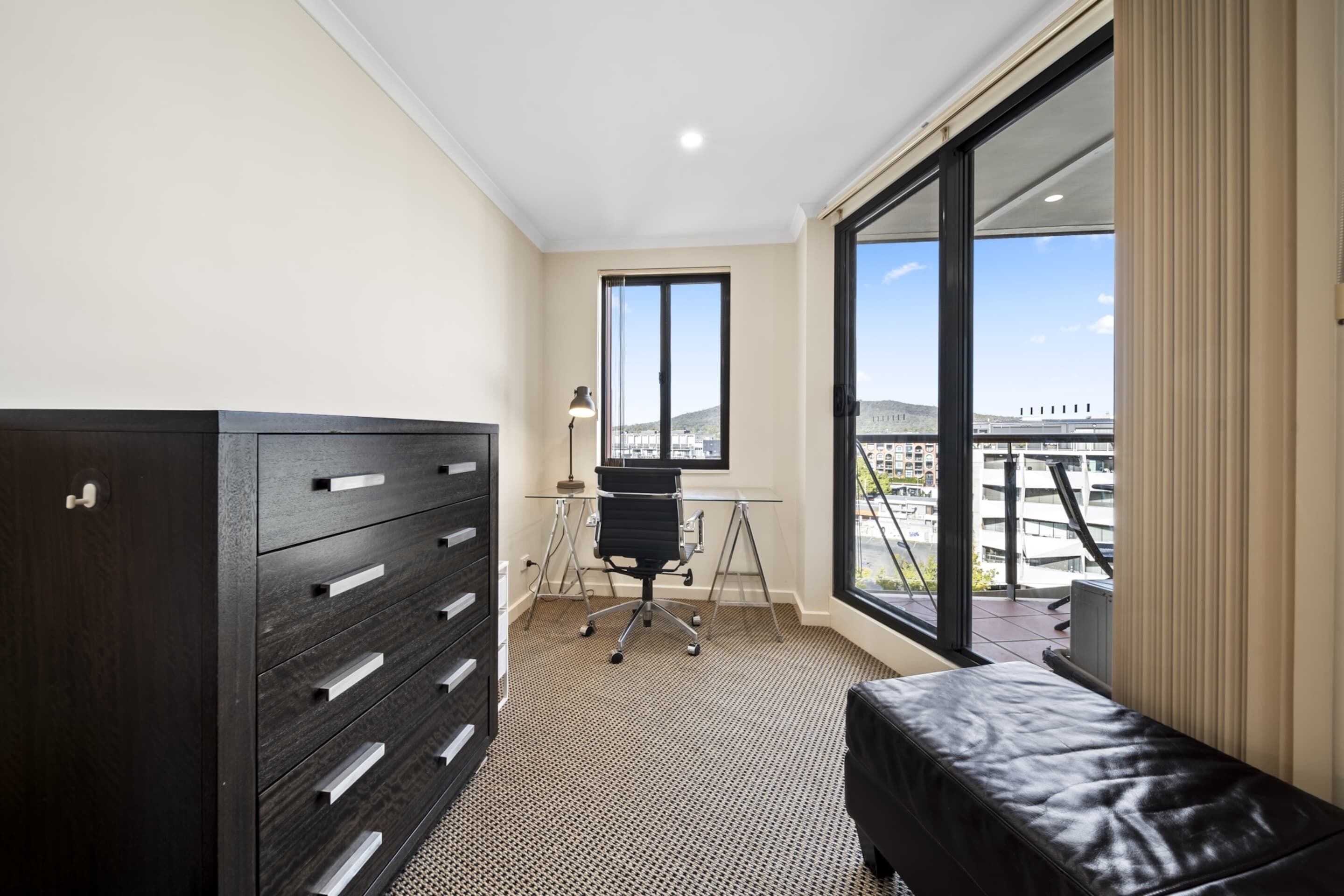 A modern room with a large window, a desk, a chair, and a dark wooden wardrobe.